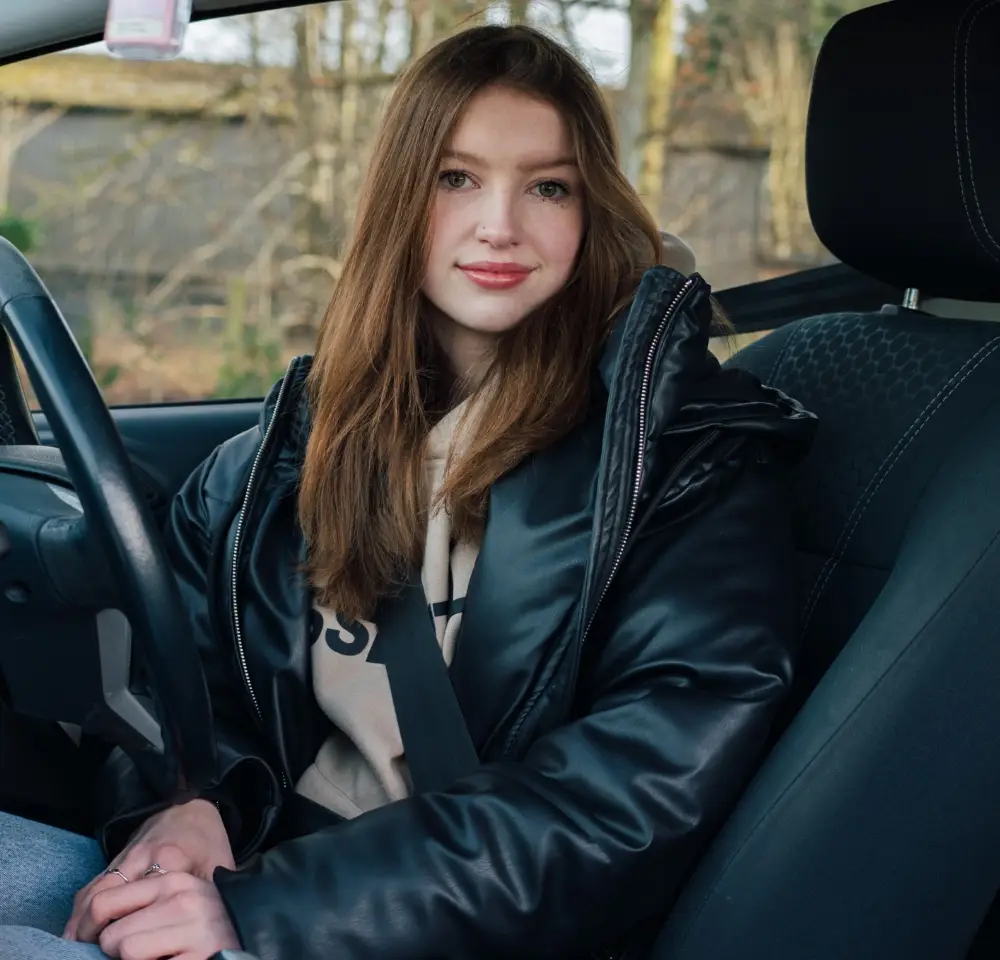 A young woman with long brown hair, wearing a black jacket and beige hoodie, sits in the driver's seat of a car, smiling gently at the camera—perhaps enjoying peace of mind with classic car insurance. Trees and a fence are visible through the window.