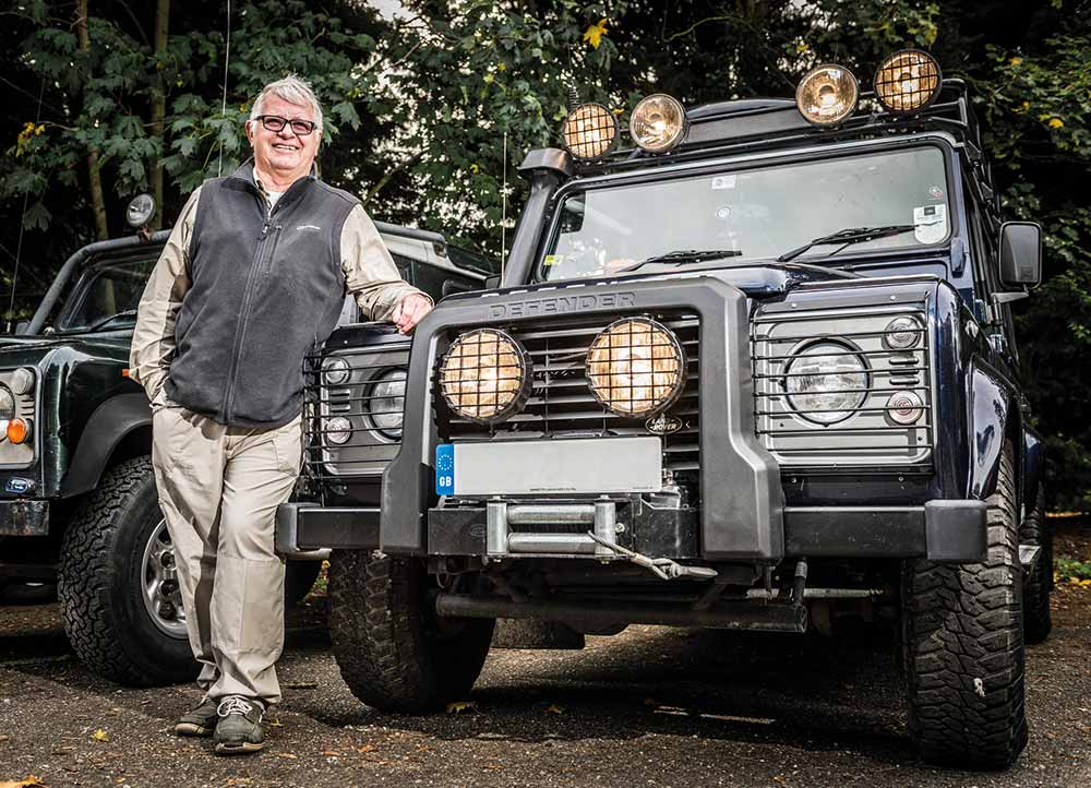 A smiling person in glasses and casual clothes stands next to a rugged black Land Rover Defender with extra lights—an ideal candidate for modified car insurance—parked outdoors near another vehicle and surrounded by trees.