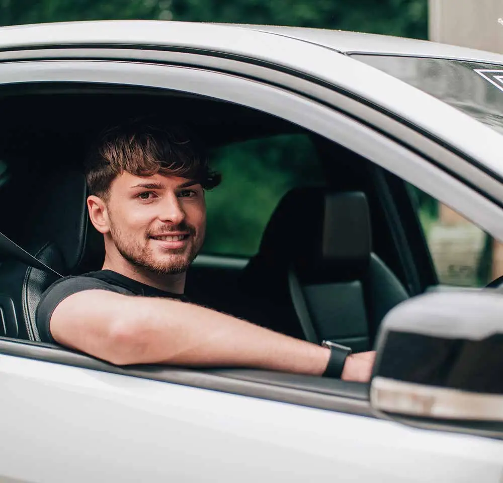 A smiling man with short brown hair and a beard sits in the driver's seat of a white car, his left arm resting on the open window, looking at the camera—perhaps considering modified car insurance. Green trees blur softly in the background.