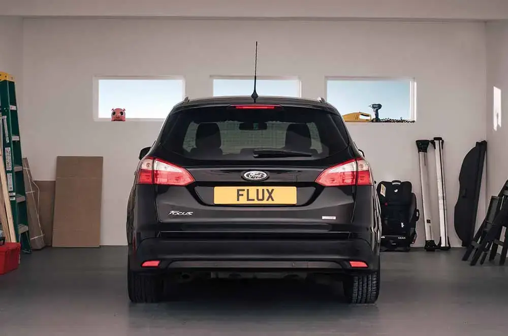 A black Ford Focus with a yellow FLUX number plate, perfect for those seeking modified car insurance, is parked inside a tidy garage. Various items, including a green ladder and tools, are stored against the white walls.