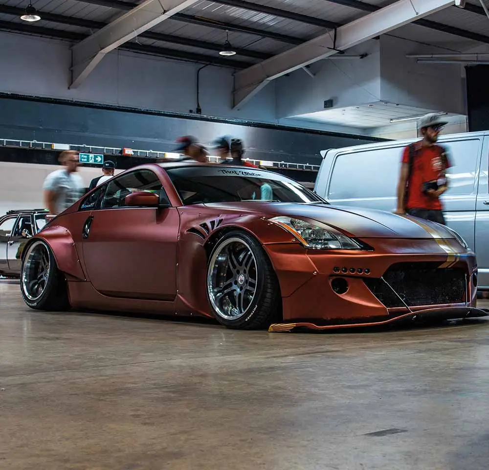 A modified bronze Nissan 350Z with wide body kit, custom wheels, and lowered suspension is displayed indoors at a car show—reminding owners to consider modified car insurance for unique builds like this. Blurred people walk nearby.