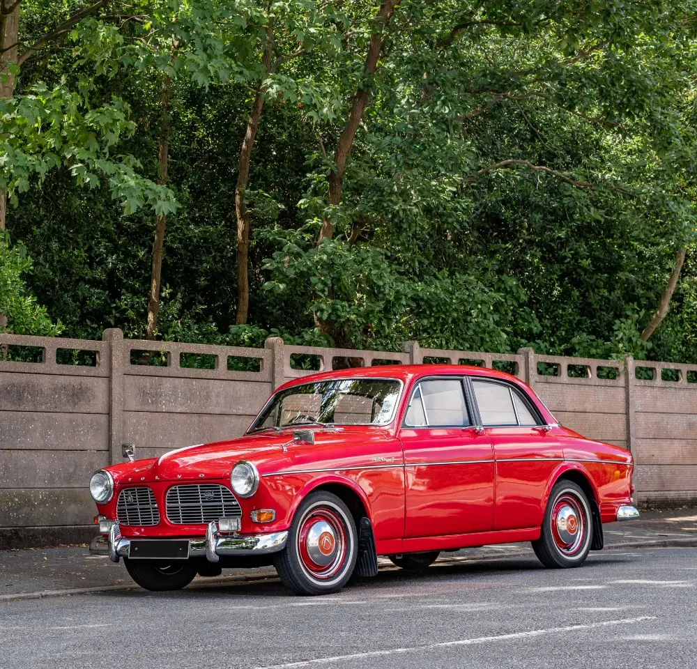 A bright red vintage car, covered by classic car insurance, is parked on a street beside a concrete fence and lush green trees on a sunny day.