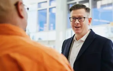 A man in a dark blazer and glasses smiles whilst chatting with another person in an orange shirt, inside a brightly lit office—perfect for team carousel photos or project teasers.
