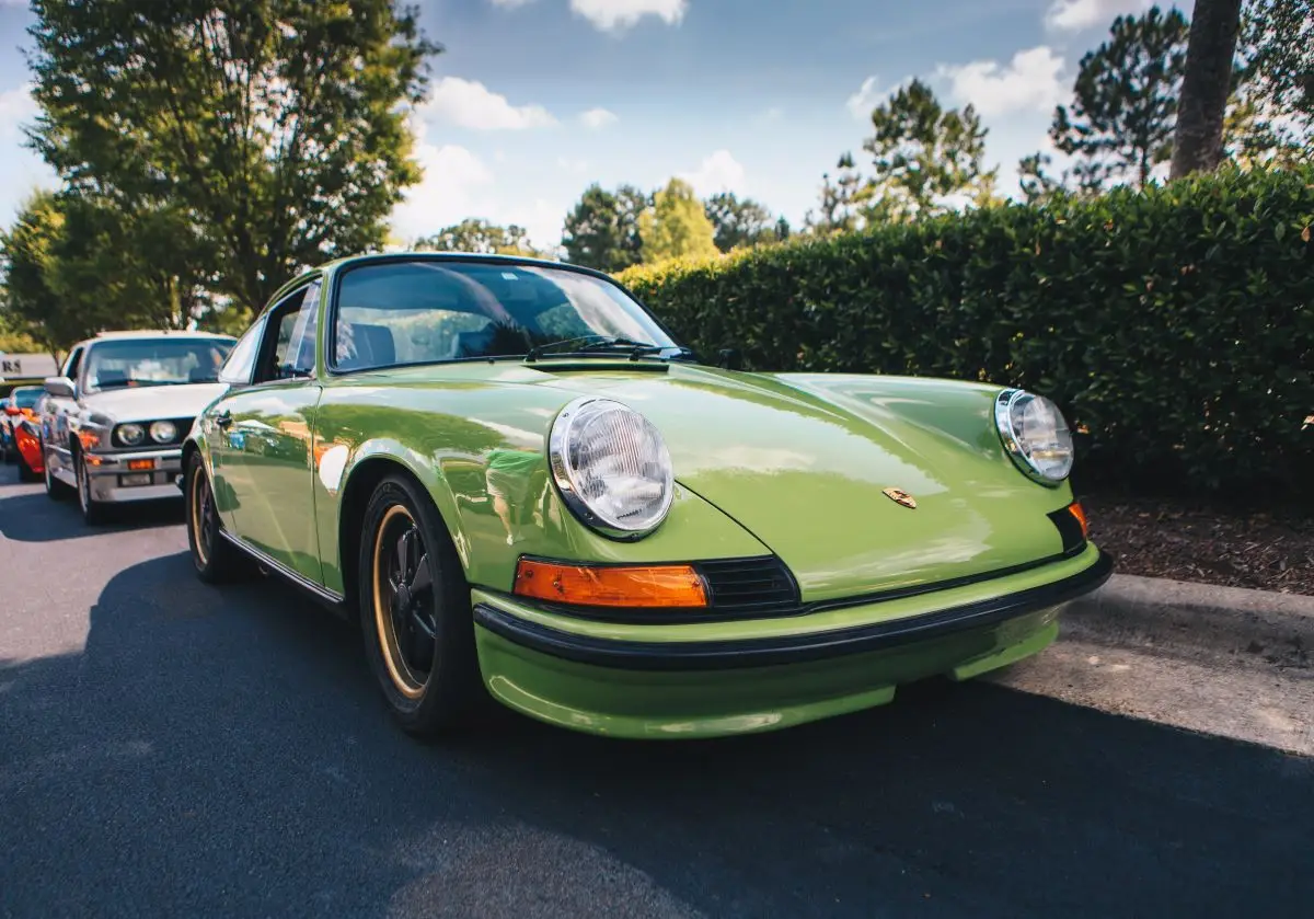 A classic green Porsche sports car, ideal for those considering modified car insurance, is parked on a road with trees and another vintage car in the background under a partly cloudy sky.