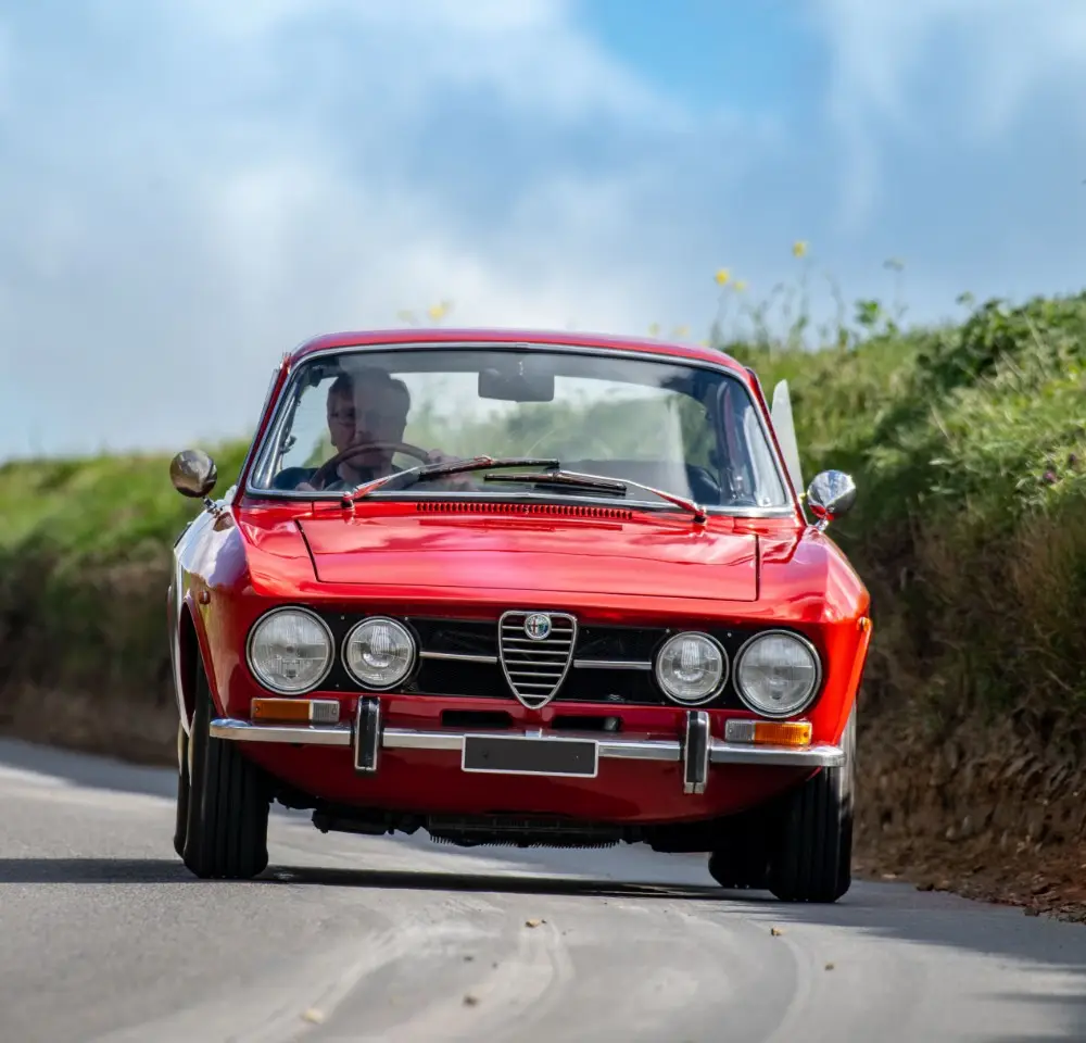 A classic red Alfa Romeo sports car drives along a rural road, green grass on either side and a blue sky in the background—a perfect drive for anyone who appreciates vintage style and values classic car insurance.