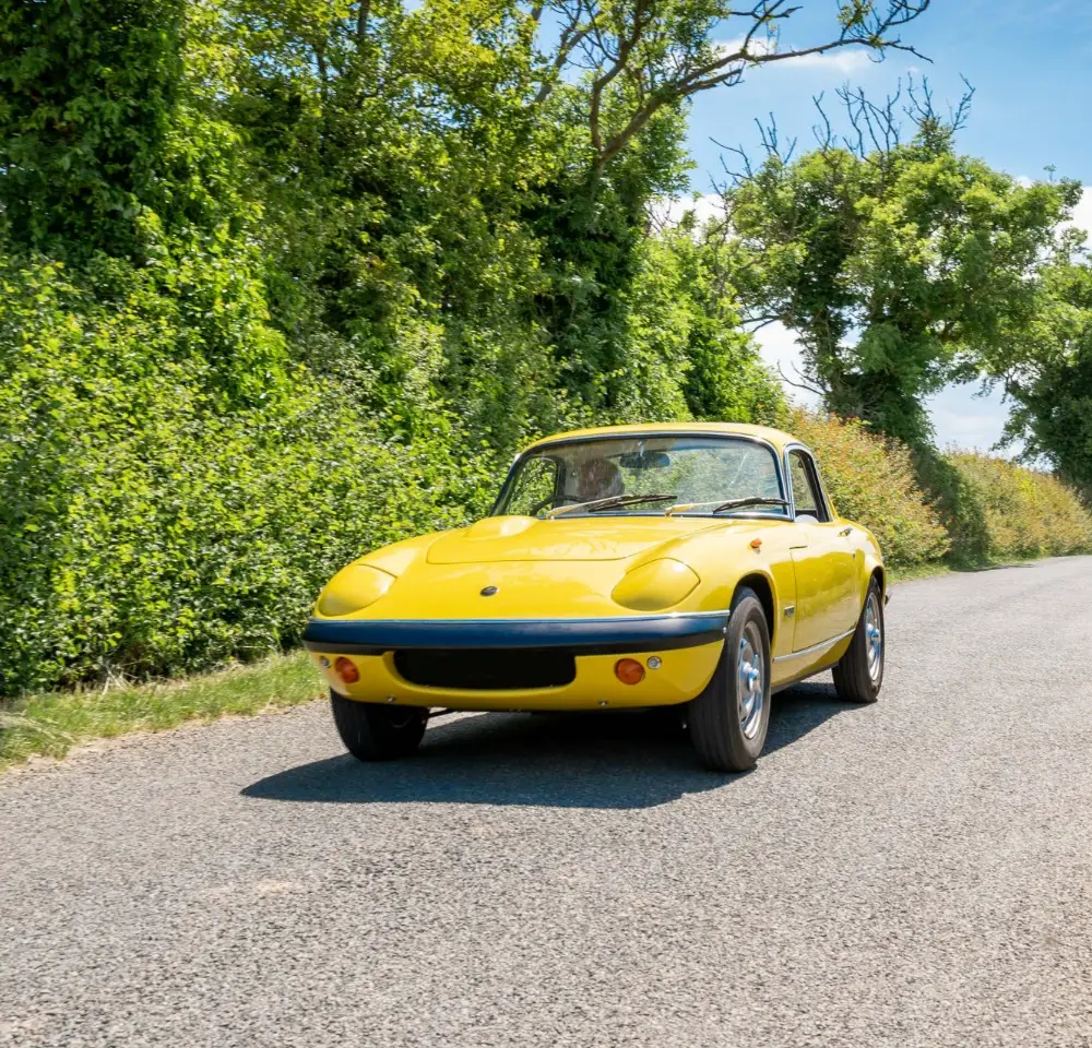 A yellow classic sports car, covered by classic car insurance, drives on a sunny rural road bordered by green trees and bushes.