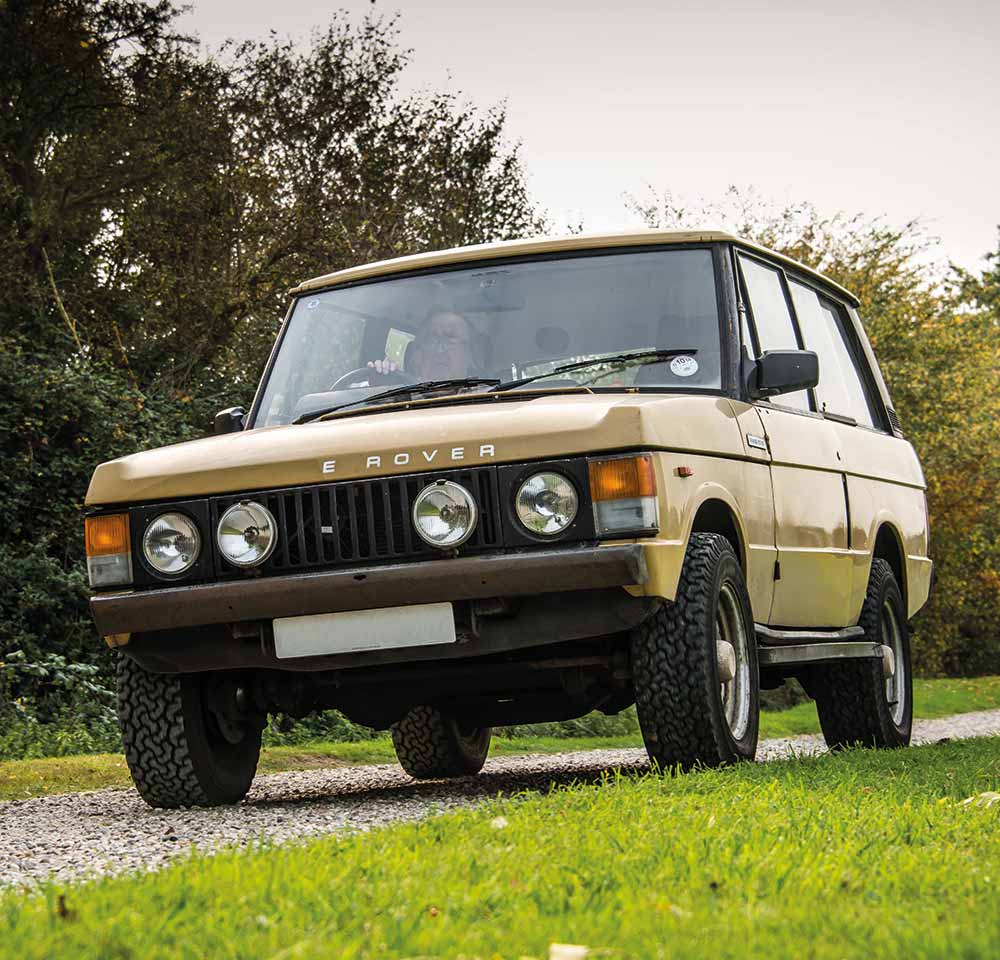 A classic beige Land Rover SUV, covered by off-road insurance, is parked on a gravel path surrounded by greenery and trees, viewed from a low front angle.