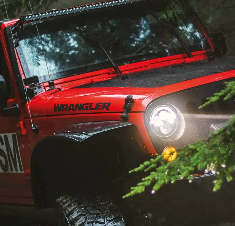 A bright red Jeep Wrangler with headlights on is parked off-road in a forest, partially obscured by green tree branches and wet from rain—an ideal scene for showcasing reliable off-road insurance.