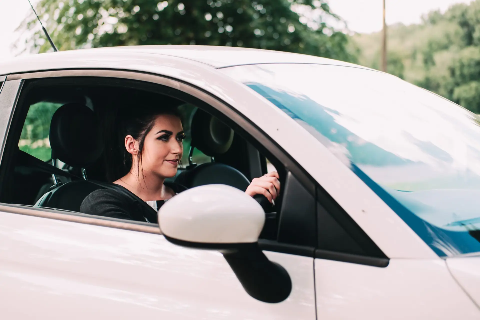 A woman with dark hair sits in the driver's seat of a white car, looking forward and holding the steering wheel, possibly benefiting from black box car insurance, with trees visible outside the window.
