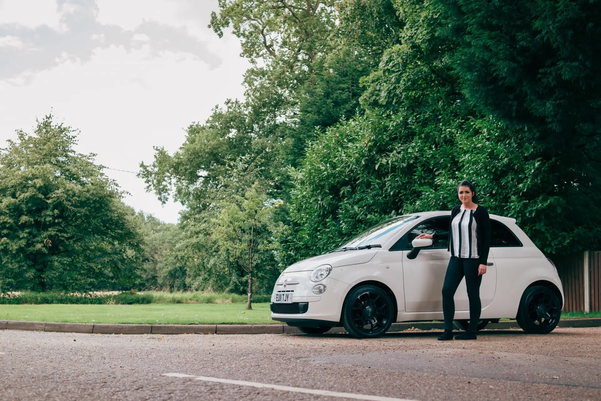 A woman in black trousers and a white-striped shirt stands next to a small white car, possibly equipped with black box car insurance, parked by the kerb amid trees and greenery on a cloudy day.