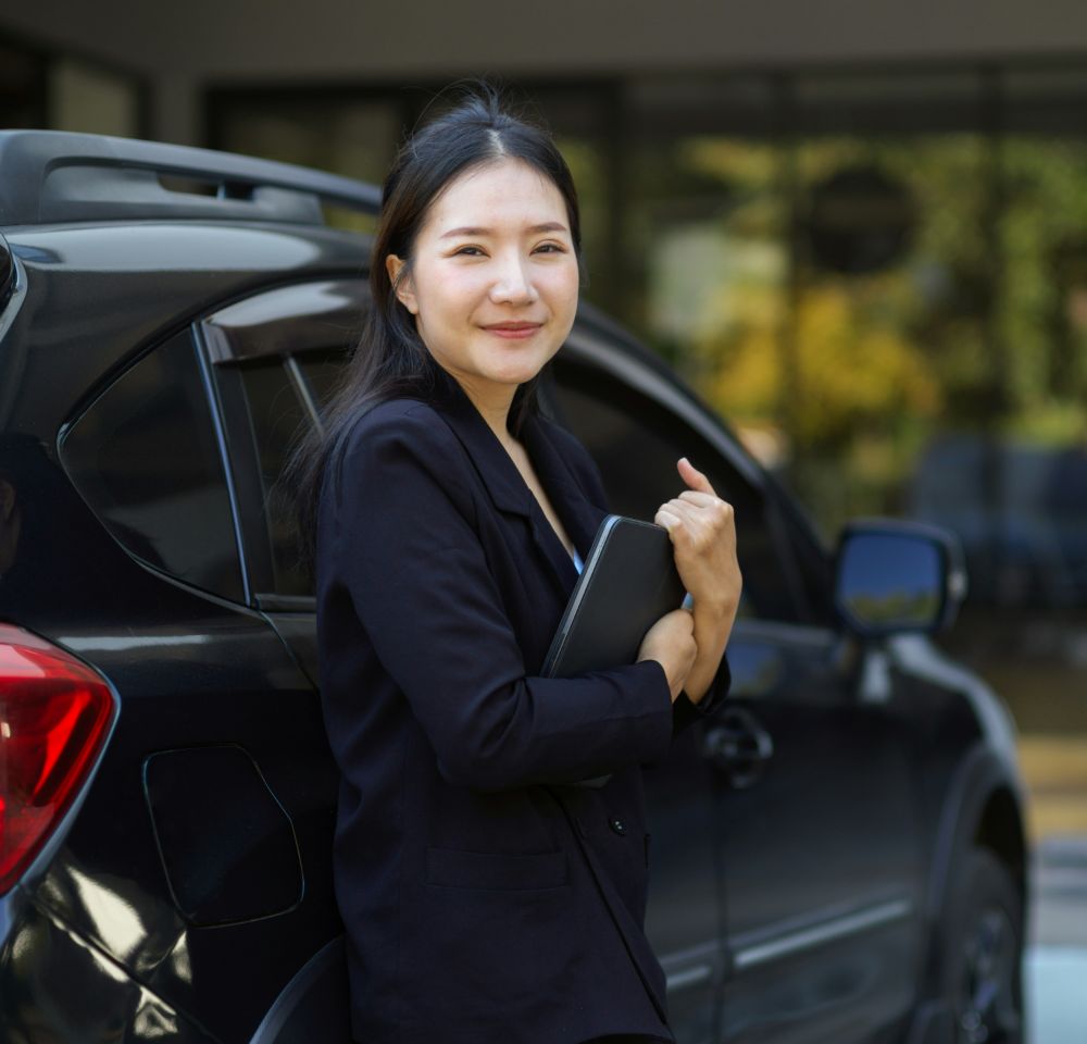 A woman in a black blazer stands beside a sleek company car, smiling and holding a tablet. The background is blurred, showing a building and greenery.