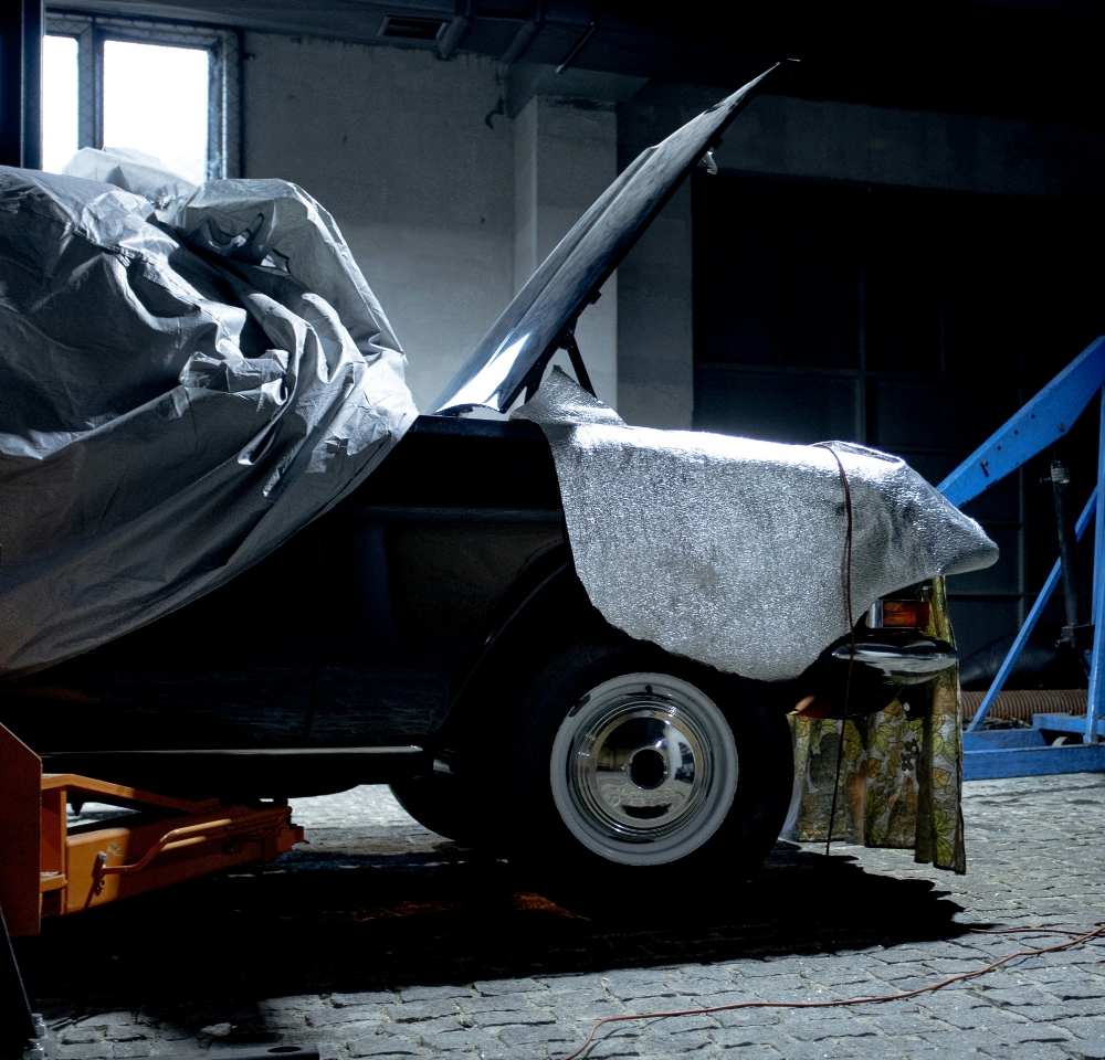 A partially covered car with its bonnet open sits on a cobblestone floor in a dimly lit garage, with tools and equipment nearby—an ideal scene for Laid-up Insurance or Build-up Cover to protect your prized vehicle during storage or restoration.