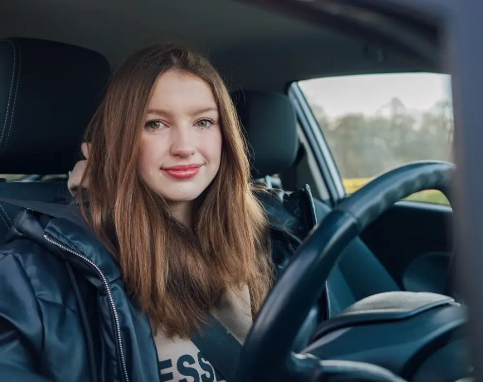 A young woman with long brown hair sits in the driver’s seat of her car, wearing a black jacket and seatbelt, smiling at the camera. Daylight streams through the windows—a confident driver ready for her black box car insurance journey.