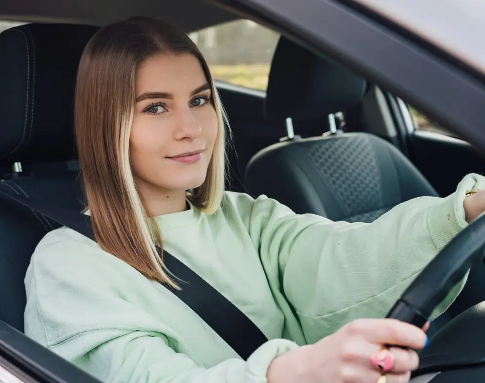 A young woman with straight blonde hair, wearing a light green jumper and seatbelt, sits in the driver's seat of a car, smiling slightly while looking towards the camera—perhaps confident thanks to her black box car insurance.