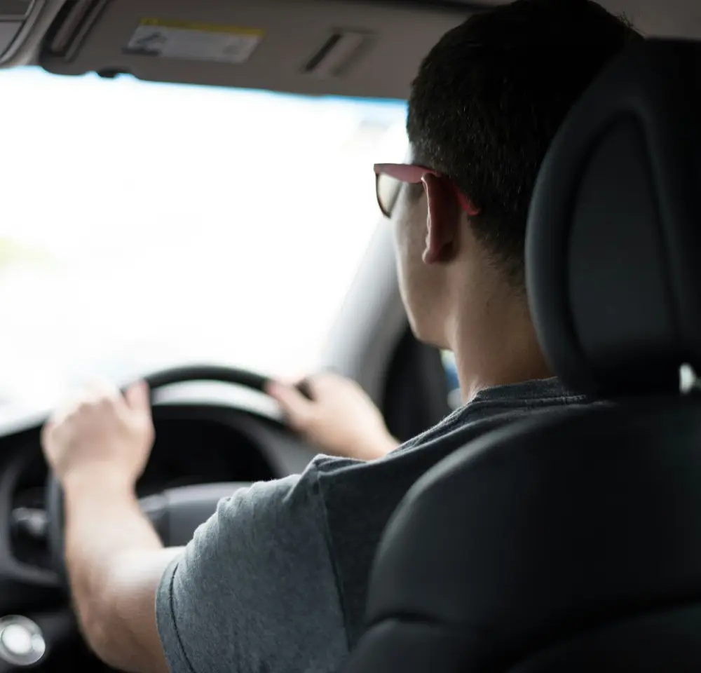 A person wearing glasses is driving a car, viewed from behind the driver’s seat. The dim interior and focused grip on the steering wheel suggest responsibility—an important factor when considering drink driving insurance.