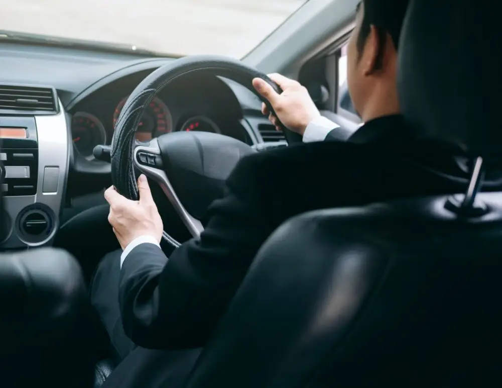 A person in a black suit is driving a car, viewed from the back seat. The driver's hands are on the steering wheel and the dashboard with gauges and car controls is visible, suggesting a professional setting possibly requiring company car insurance.