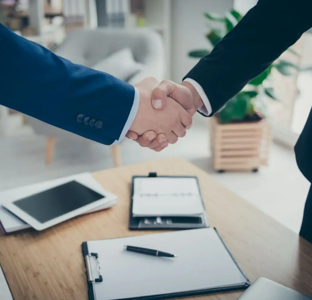 Two people in business suits shake hands over a desk with a clipboard, pen, and tablet, suggesting a professional agreement or business meeting in a modern office setting—possibly discussing company car insurance options.