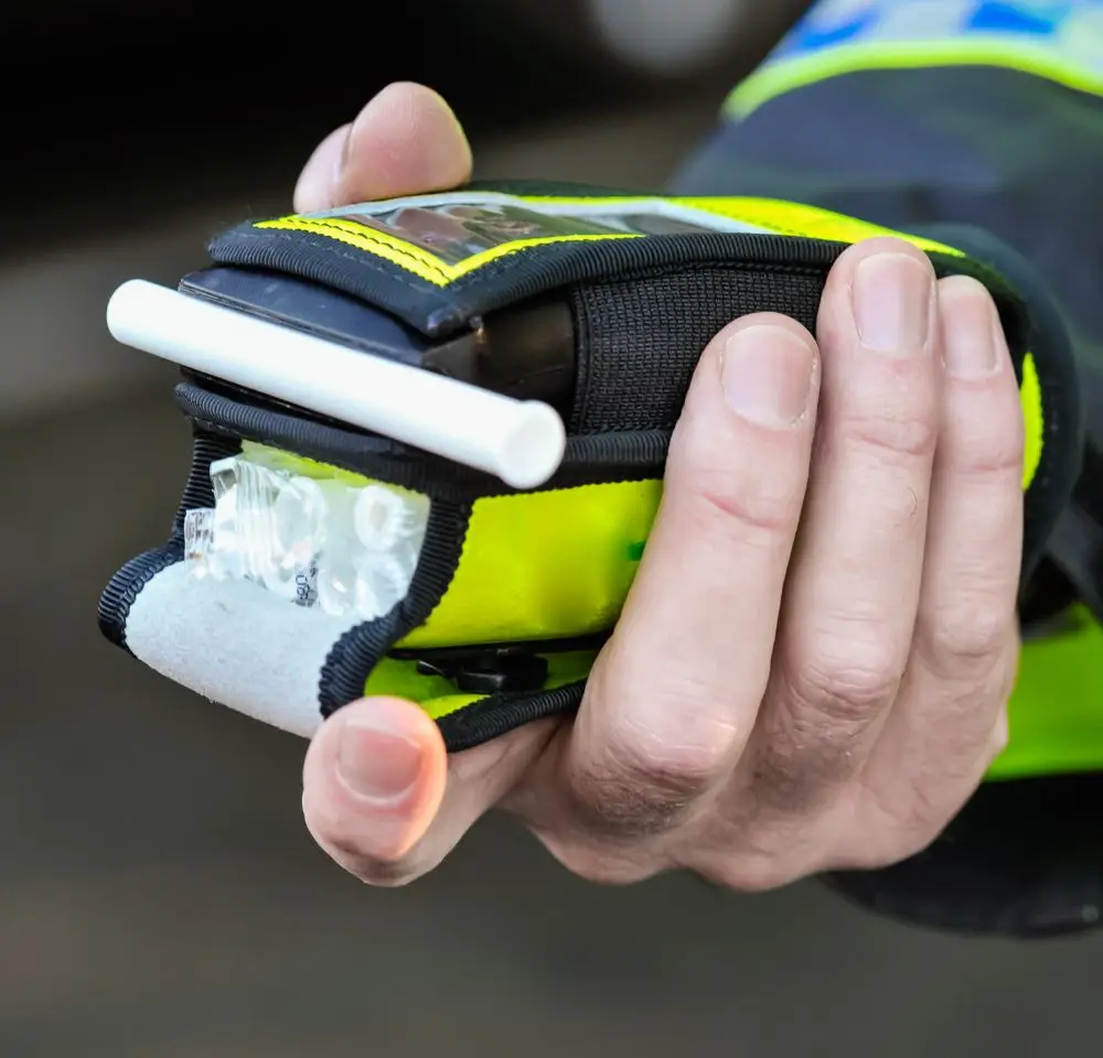 A person holds a portable breathalyser device with a white mouthpiece, used for measuring blood alcohol content, often essential for those seeking DG10 conviction insurance, against a blurred background.