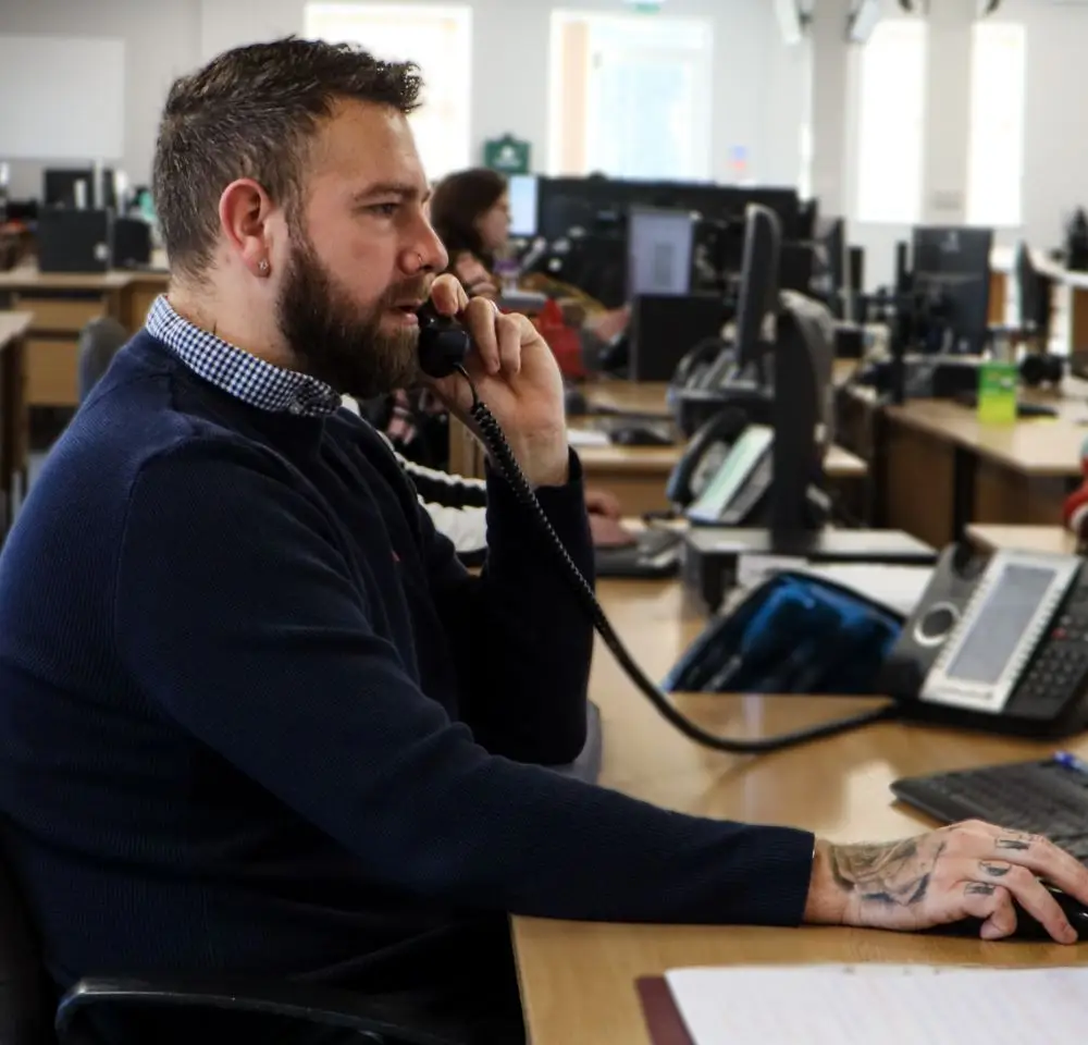 A bearded man with tattoos on his hand talks on a landline phone at an office desk, surrounded by paperwork and computer equipment—possibly discussing DG40 insurance. Other desks and people are visible in the background.
