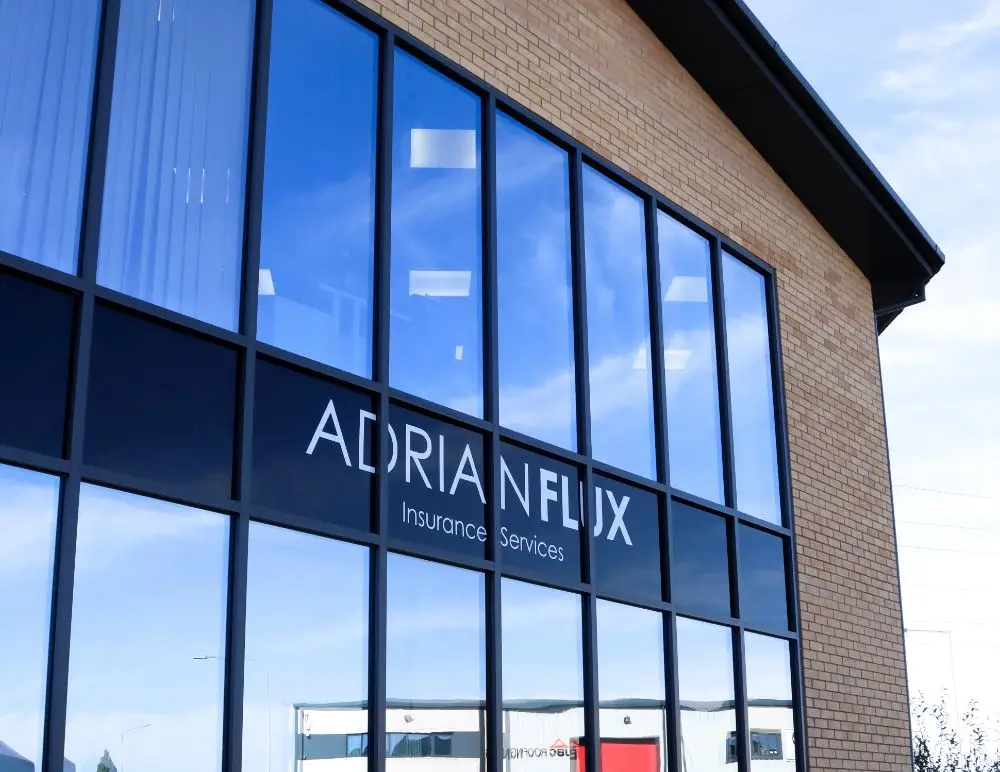 A modern brick building with large reflective windows displays the Adrian Flux Insurance Services logo, known for helping clients with a DR20 driving conviction. The blue sky and clouds are mirrored in the glass.