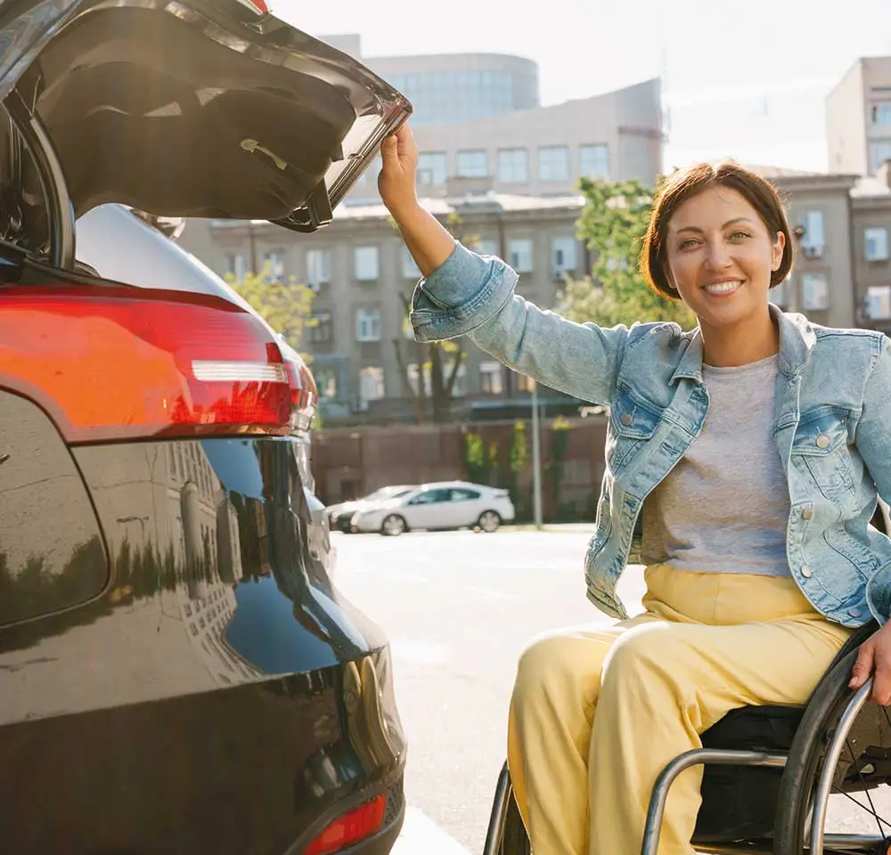 A woman in a wheelchair smiles whilst opening the boot of a black car, enjoying a sunny day in the city—highlighting the independence offered by Disabled Driver Insurance.