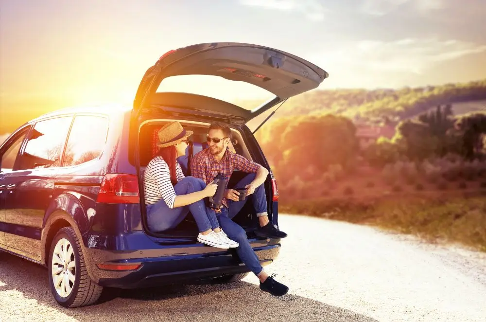Two people sit in the open boot of a parked car on a sunny day, smiling and talking, with a scenic landscape and sunset in the background.