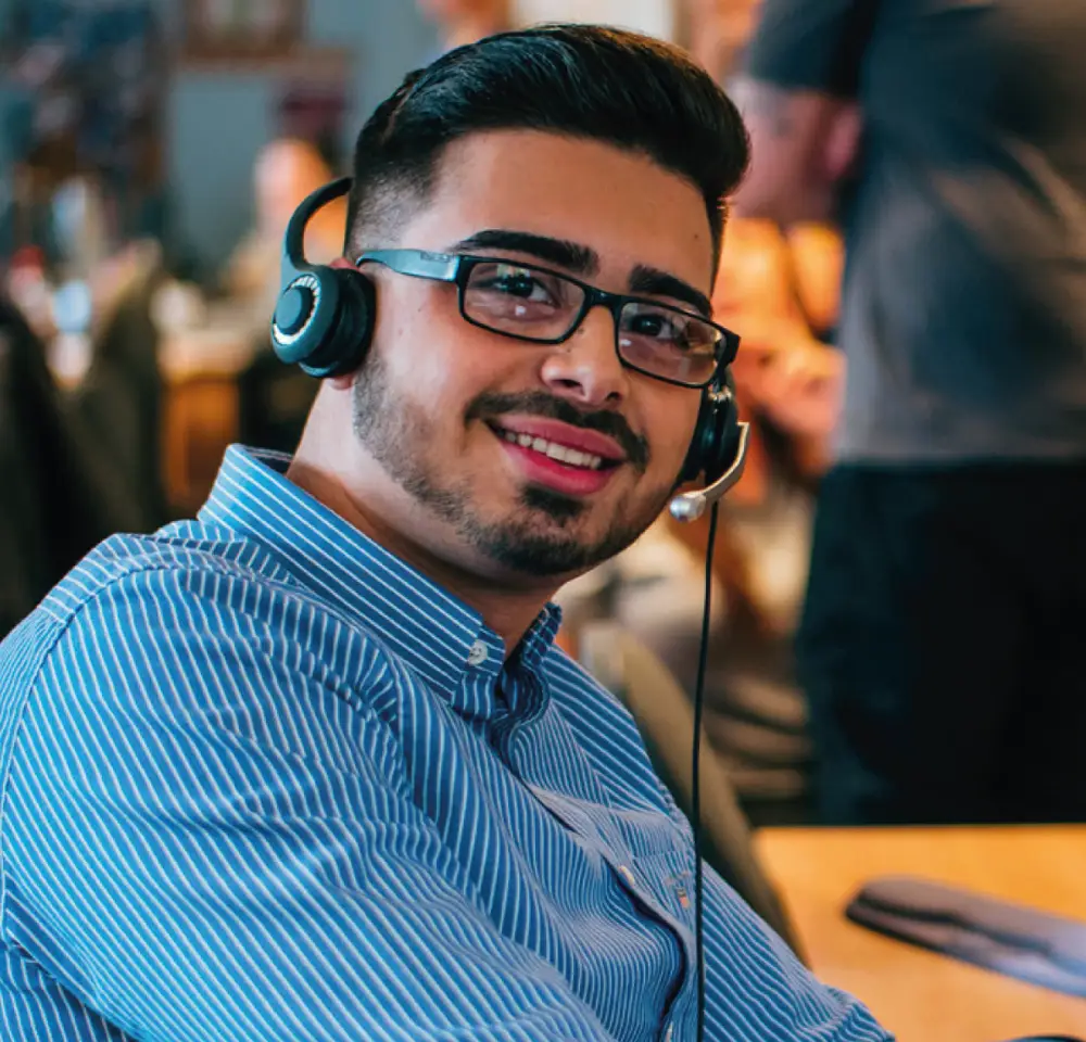 A young man wearing glasses and a striped blue shirt smiles at the camera whilst sitting at a desk with a headset, ready to assist customers seeking drink-driving insurance in a busy office environment.