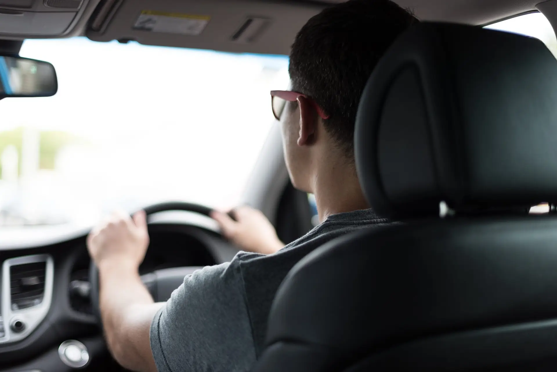 A person wearing spectacles is driving a car, viewed from the back seat. The driver with short hair has both hands on the steering wheel, while the interior—suggestive of a black box car insurance set-up—is clearly in focus.
