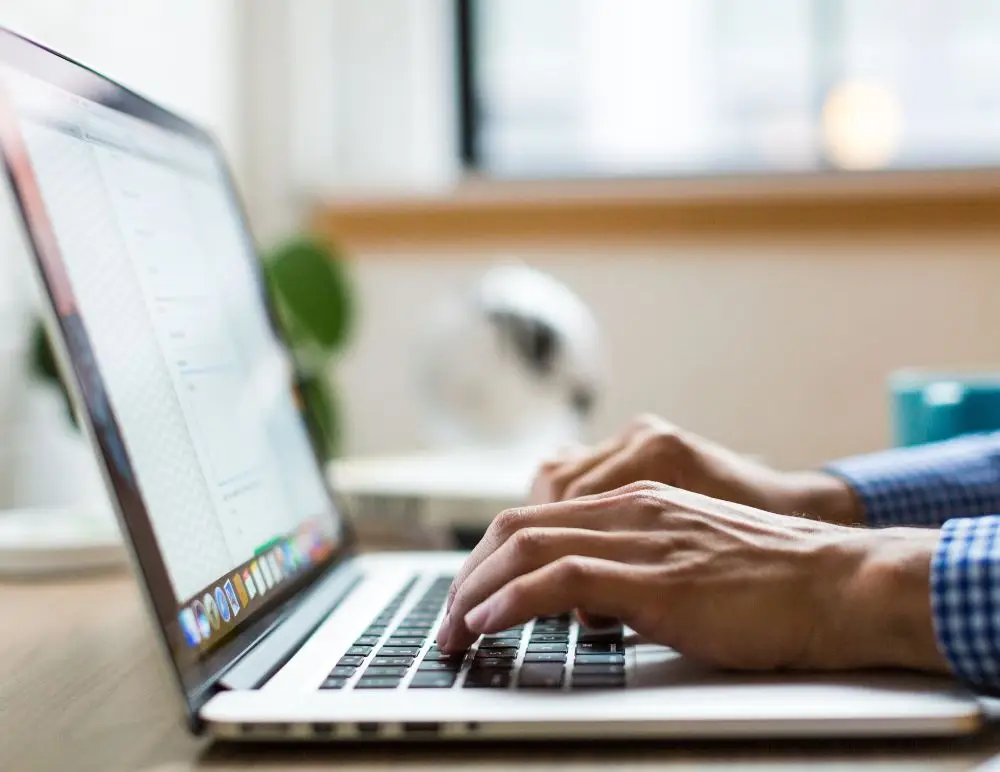 Close-up of a person’s hands typing on a laptop keyboard at a desk, possibly researching car insurance or cancelled policies, with a blurred background including a window and some office items. The person is wearing a blue checked shirt.