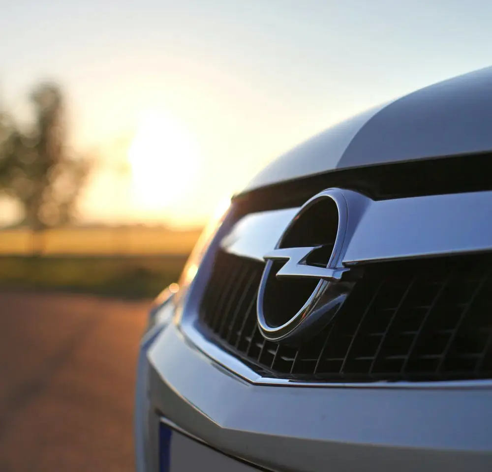 Close-up of the front grille and logo of an Opel car, with a blurred background of a road and trees at sunset—a reminder to always keep your car insurance up to date for peace of mind on every journey.