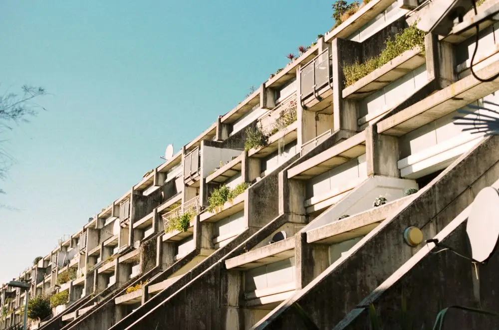 A concrete block of flats with staggered balconies and greenery under a clear blue sky, viewed from a low angle. The geometric, modern design highlights visible railings and satellite dishes—ideal for those seeking comprehensive HMO landlord insurance.