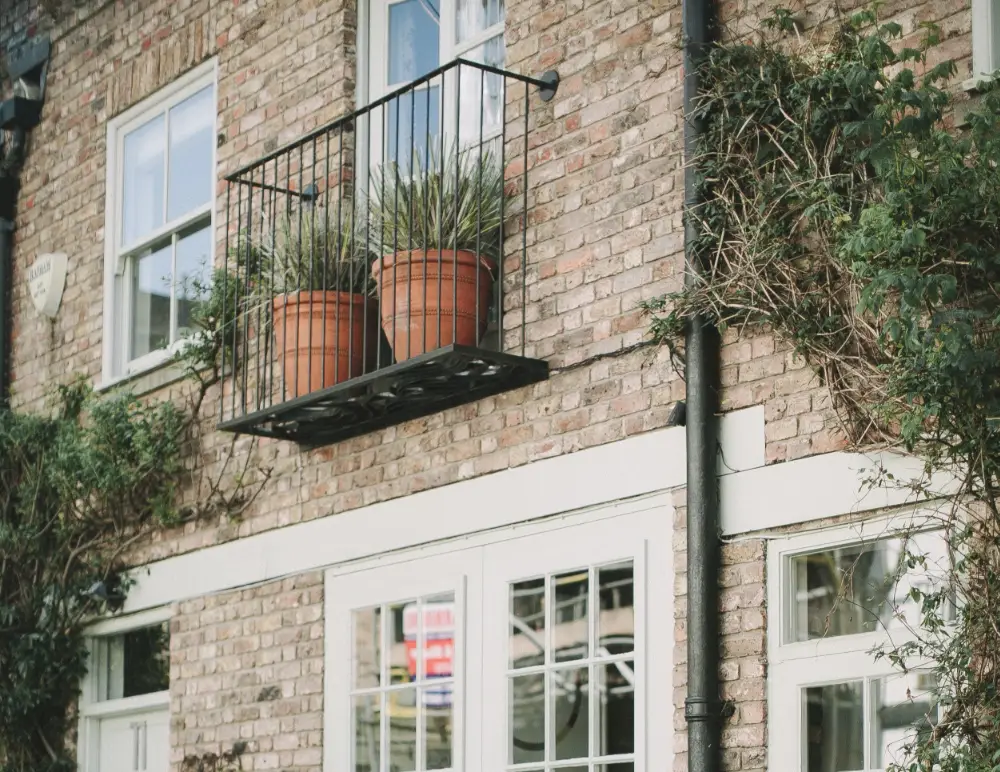 A small black metal balcony with two potted plants is attached to the brick exterior of a building, above white-framed windows and surrounded by climbing green vines—ideal for those considering converted property insurance.