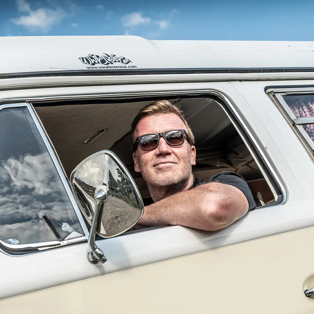 A man wearing sunglasses leans out of the window of a vintage white van, looking relaxed under a blue sky with scattered clouds—enjoying the freedom that comes with campervan insurance.
