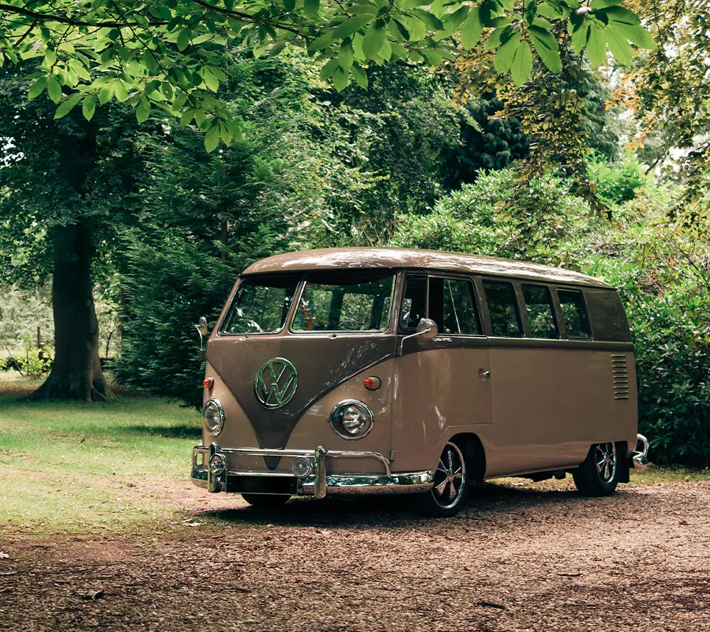 A vintage beige Volkswagen camper van, protected with Campervan Insurance, is parked on a dirt track surrounded by lush green trees and grass in a peaceful, wooded area.