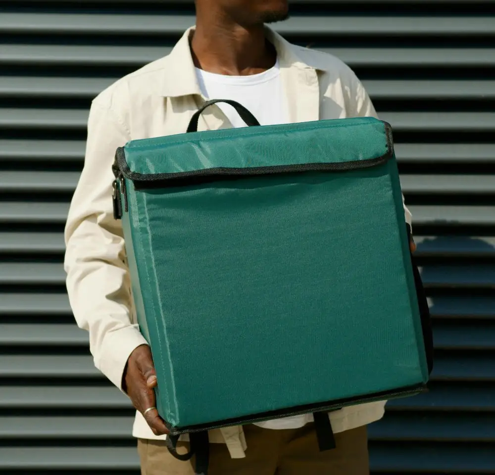 A person wearing a light jacket and white shirt holds a large green insulated delivery bag in front of their chest, standing against a dark horizontal striped background—an image reflecting the importance of courier van insurance for safe deliveries.