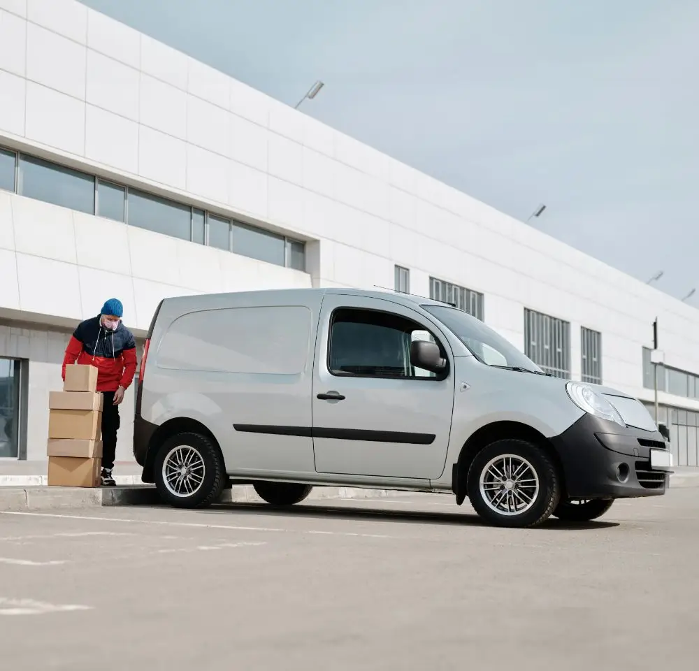 A delivery worker in a uniform stands next to a white van, stacking cardboard boxes outside a modern industrial building on a cloudy day, highlighting the importance of courier van insurance for secure and reliable service.