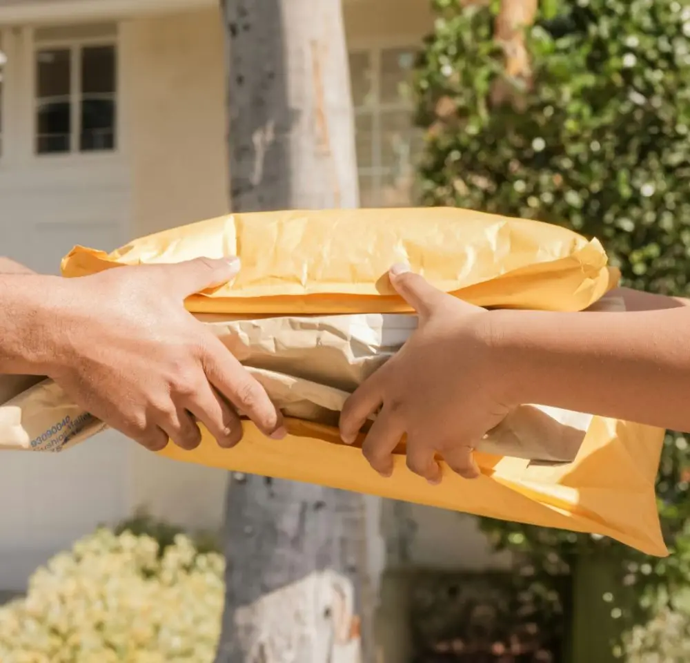 Two people's hands exchange several parcels wrapped in brown and yellow envelopes outdoors, with greenery, a building, and a courier van insurance vehicle in the background.