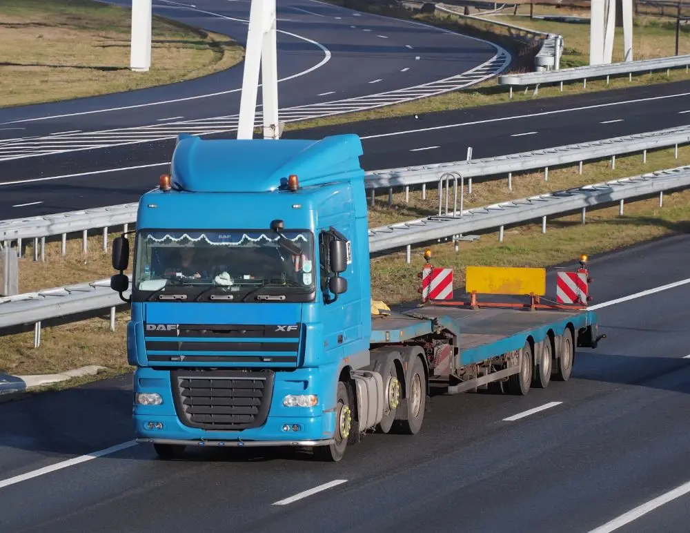 A blue lorry with an empty flatbed trailer, covered by Flatbed Lorry Insurance, drives on a multi-lane motorway bordered by metal crash barriers and grassy areas.