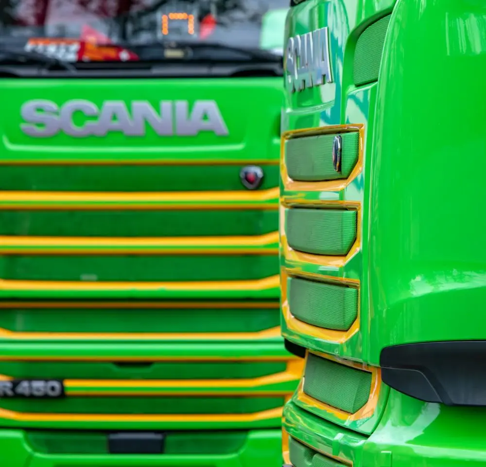 Close-up of two bright green Scania lorries with yellow accents, highlighting their front grilles and the Scania logo—ideal vehicles for those seeking Flatbed Lorry Insurance. One lorry faces forward while the other sits at an angle in the foreground.
