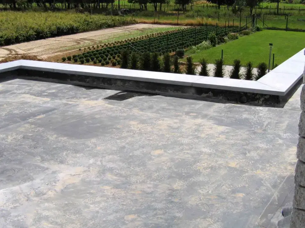 A rooftop with a concrete surface, ideal for flat roof insurance, overlooks a neatly landscaped garden featuring rows of shrubs, green grass, and a path lined with small trees.
