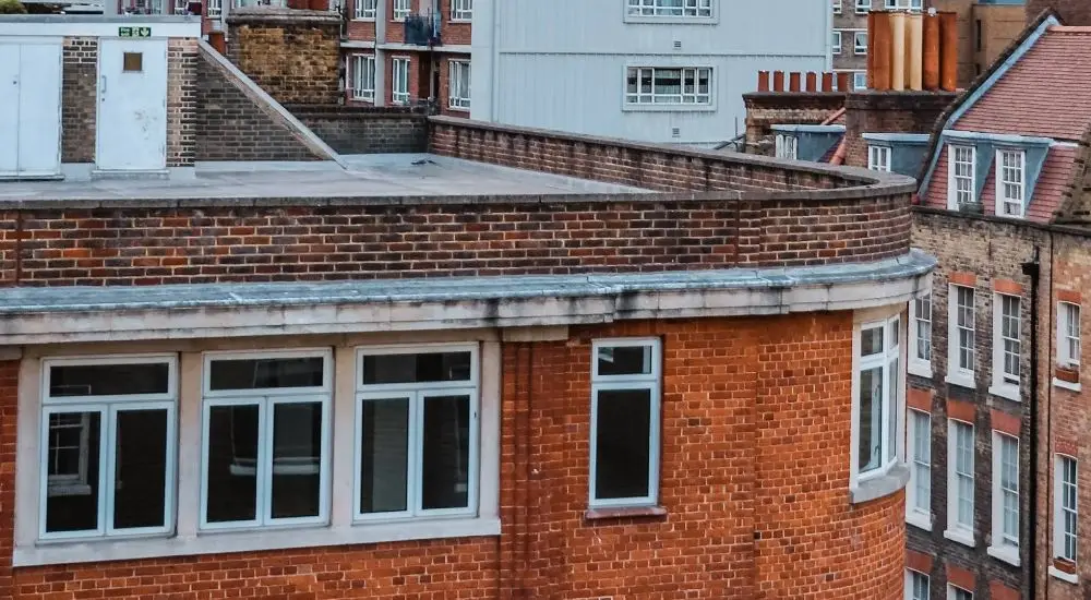 A close-up view of a curved corner of a red-brick building with white-framed windows and a flat roof, highlighting structures that may require Flat Roof Insurance in an urban setting with other brick and concrete buildings.