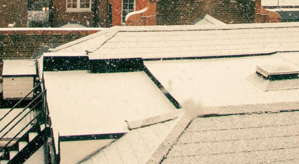 Snow falling gently onto flat rooftops, covering them with a thin, white layer. Surrounding brick buildings and small windows are visible in the background—a calm, wintry scene that highlights the importance of flat roof insurance.