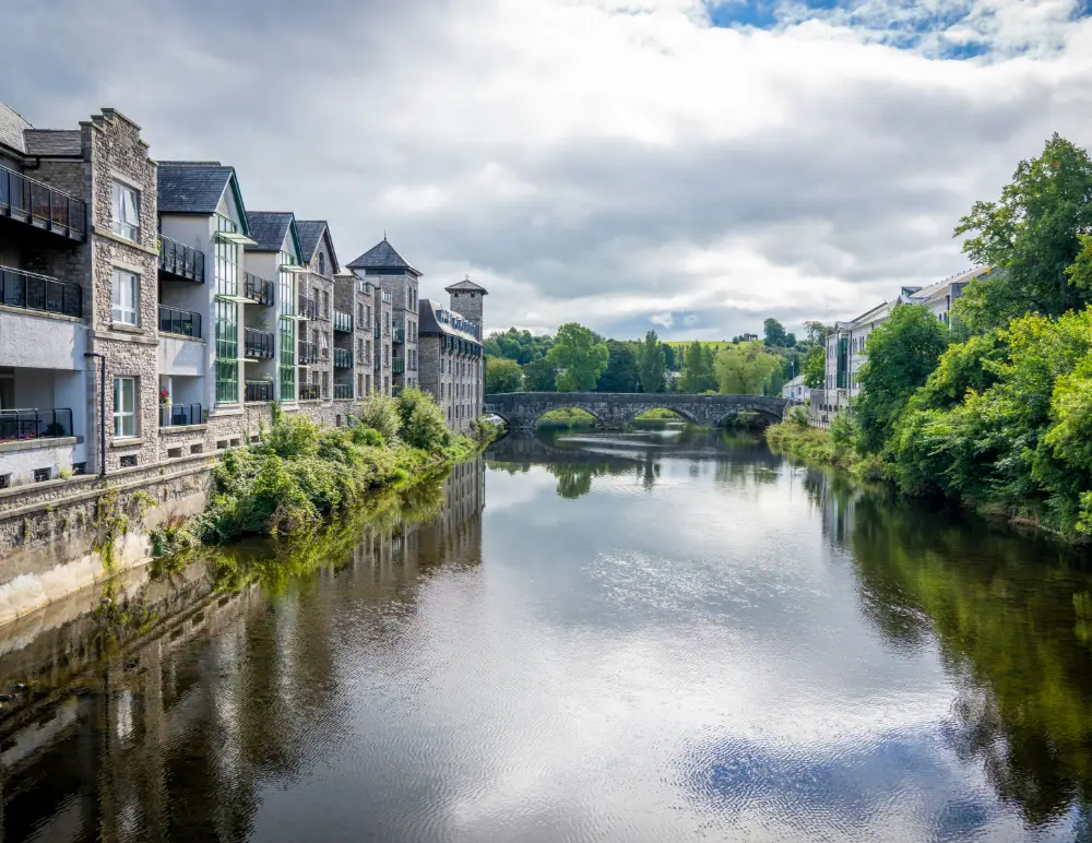 A calm river runs through a town with stone buildings on the left and trees on both sides, leading to an old arched stone bridge under a cloudy sky—a scene where understanding flood risk is essential for homeowners.