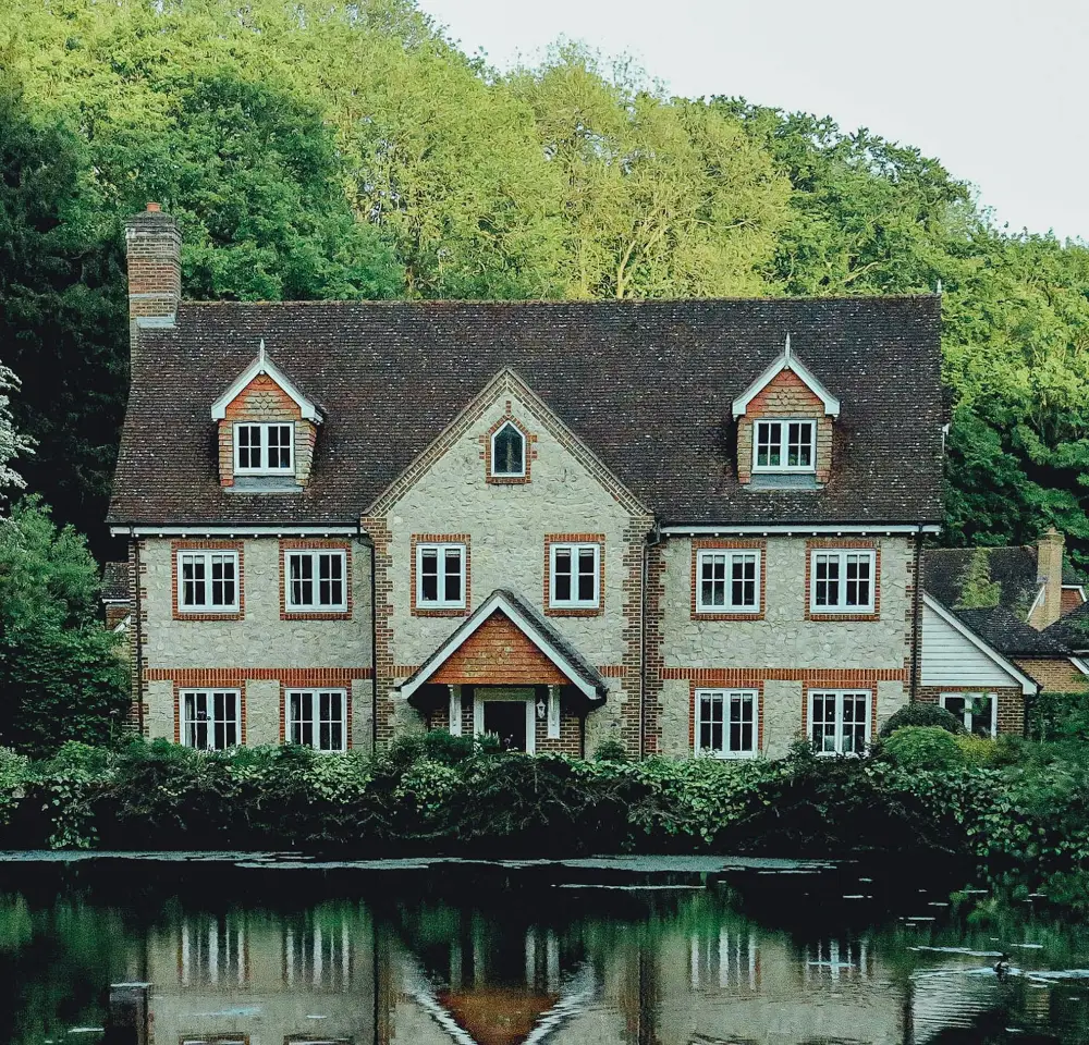 A large, two-storey brick house with three gabled windows stands before a lush green forest, its reflection shimmering in a calm pond—an idyllic setting where considering flood insurance can help protect your home from unexpected flood risks.