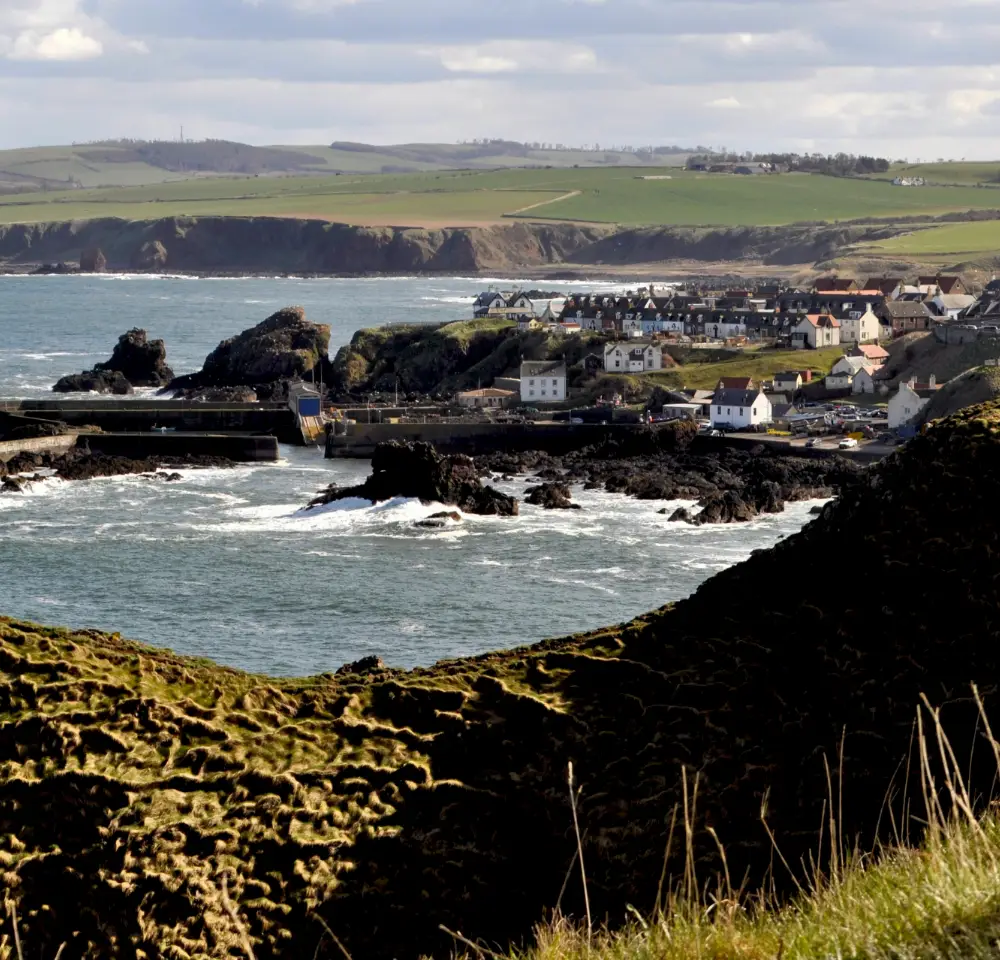 A coastal village with white houses sits by rocky cliffs and a small harbour, overlooking choppy sea waves—an ideal place to consider Flood Insurance for homes near the rolling green fields and distant hills under a partly cloudy sky.