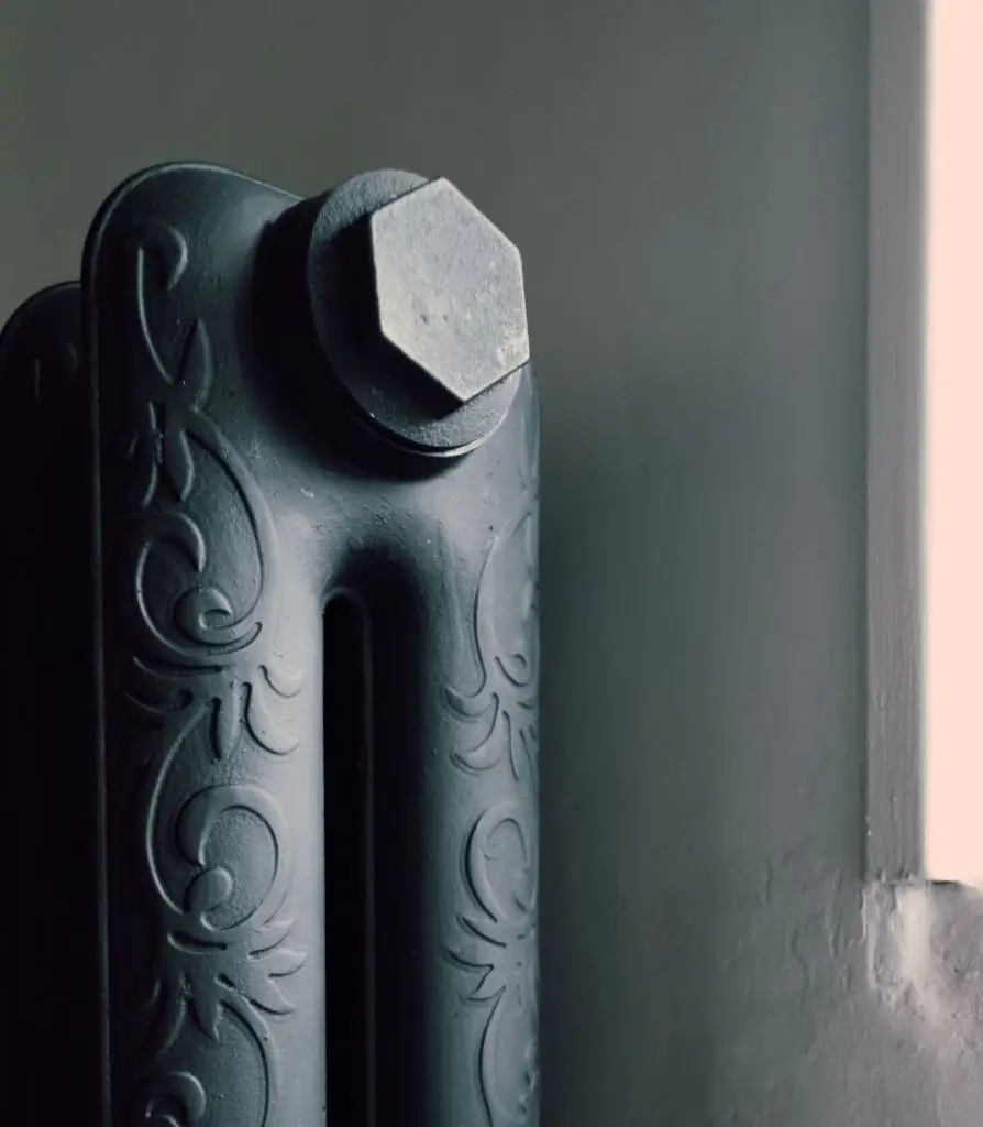 Close-up of a dark metal radiator with ornate floral patterns, positioned next to a light grey wall and illuminated by natural light from a nearby window—ideal for spaces covered by Listed Building Insurance.
