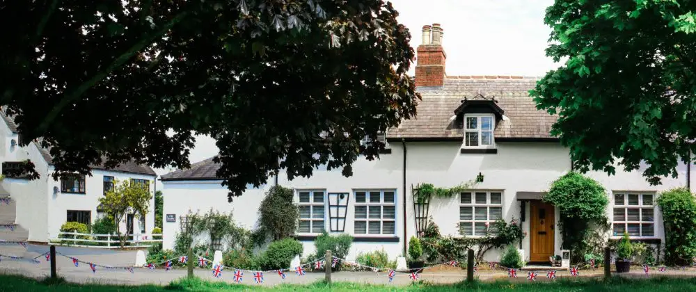 A white cottage with a tiled roof and brick chimney, covered by Listed Building Insurance, is decorated with small Union Jack flags across the front garden. The house is surrounded by green trees and shrubs on a sunny day.