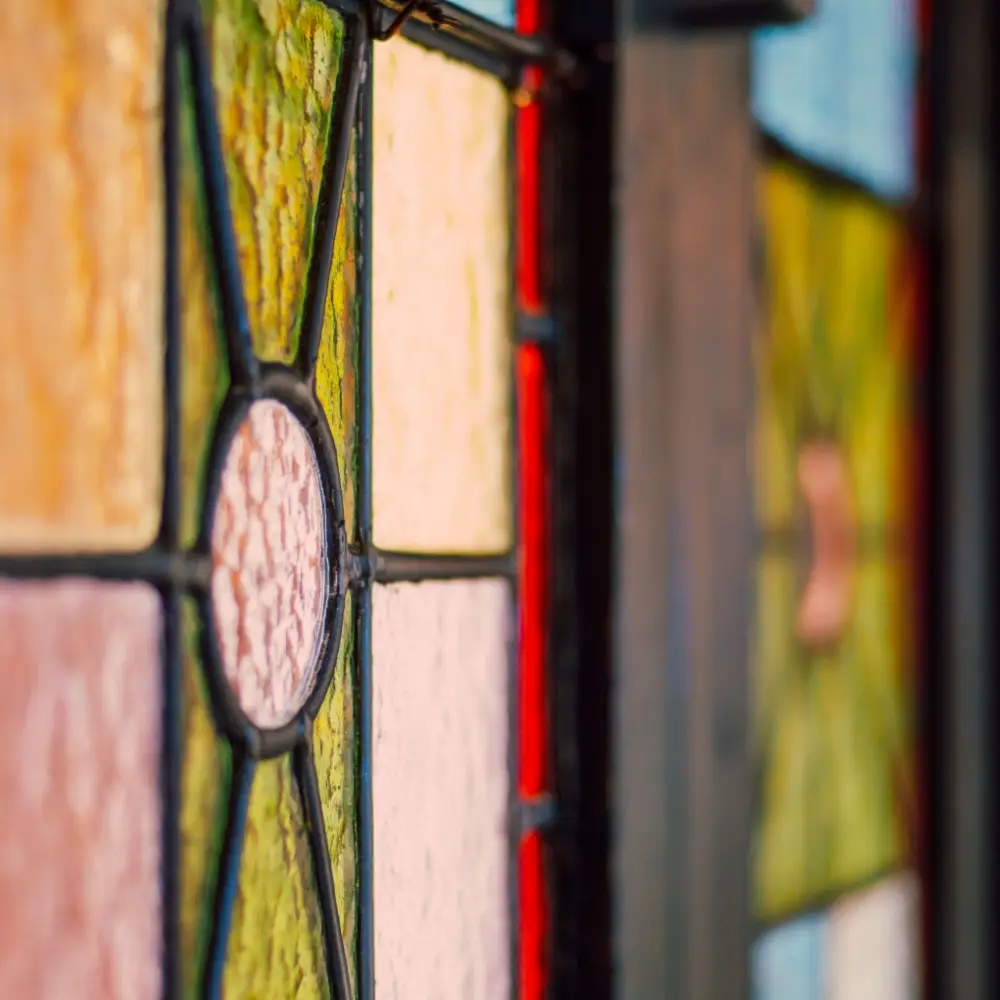 Close-up of a colourful stained glass window with textured panels in yellow, pink, and green, bordered by dark metal lines. The blurred background highlights the geometric pattern—an exquisite feature often protected under Listed Building Insurance.
