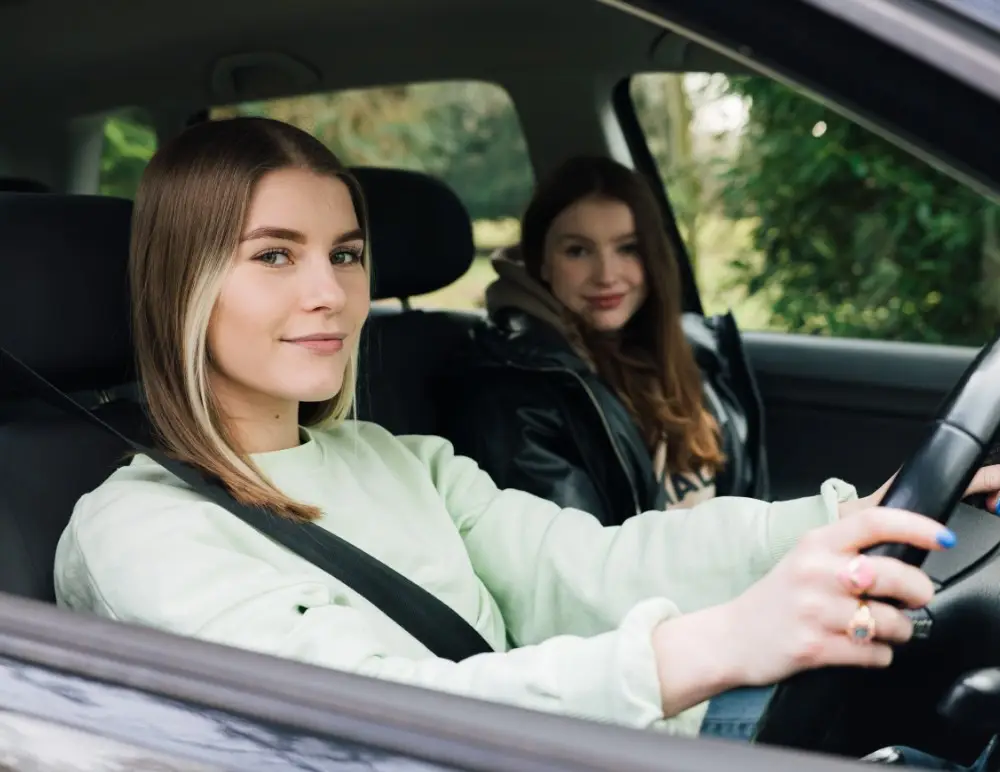 Two young women sit in a car. The driver, wearing a light green jumper, smiles at the camera—enjoying her ride with Young Driver Insurance—while the passenger grins beside her. Trees are visible outside the car windows.