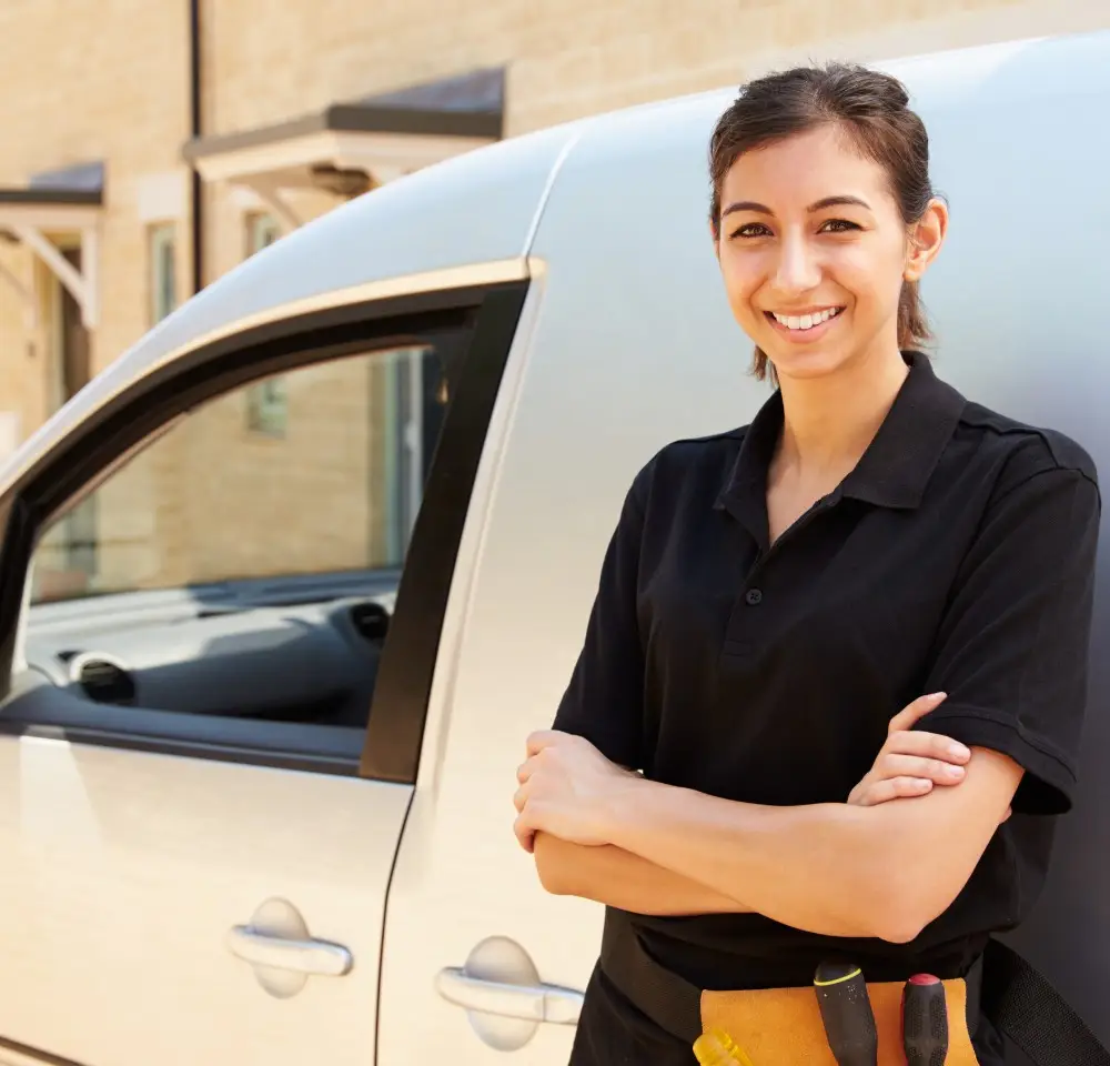 A smiling woman wearing a black polo shirt and tool belt stands with arms folded next to a white van, outdoors in a residential area, confidently knowing her vehicle is covered by van insurance.